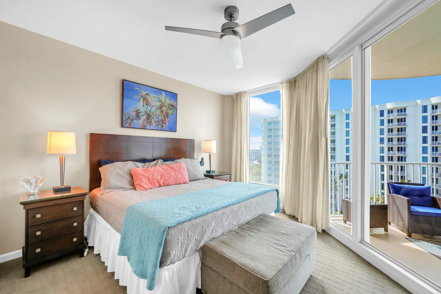 Bedroom with a wooden headboard, bedside tables with lamps, a ceiling fan, and large windows and sliding glass doors leading to a balcony.