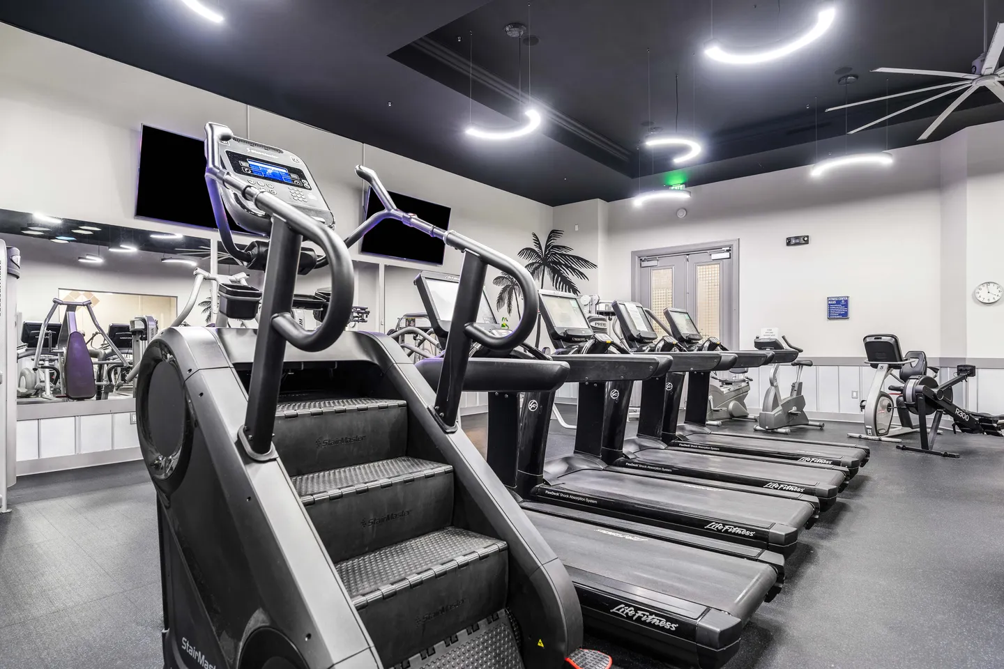 Indoor fitness center featuring a StairMaster, a row of treadmills, and curved ceiling lights.
