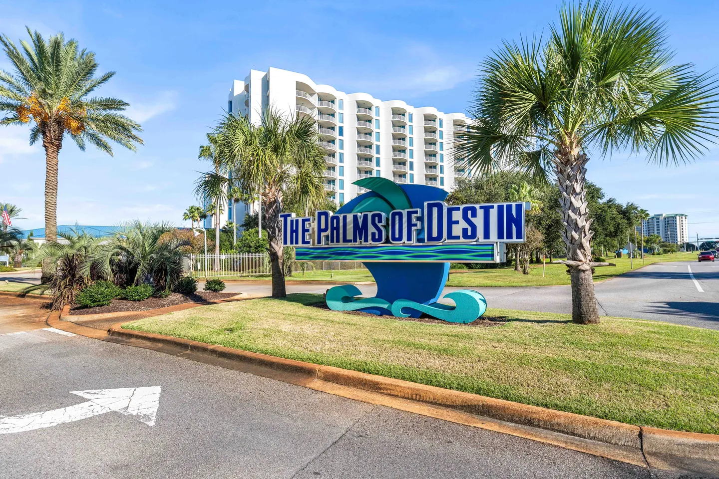 Entrance sign for The Palms of Destin framed by palm trees with a multi-story resort building in the background.