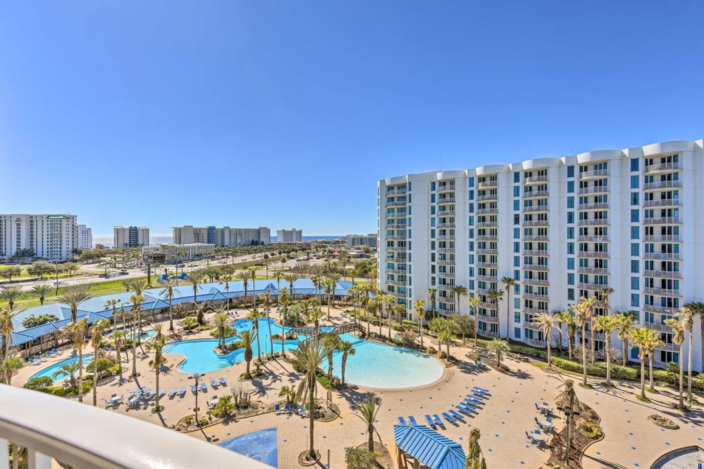Elevated view from a balcony overlooking a large resort swimming pool complex and multi-story condominium building under a clear blue sky.