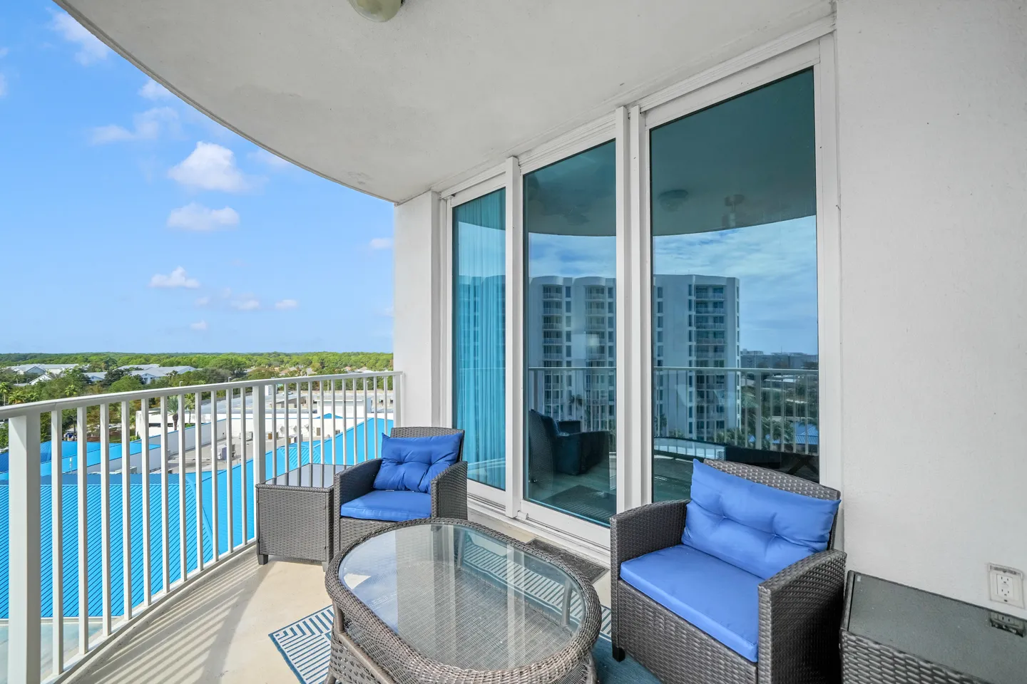Covered balcony featuring wicker armchairs with blue cushions, a glass-topped coffee table, and sliding glass doors under a bright blue sky.