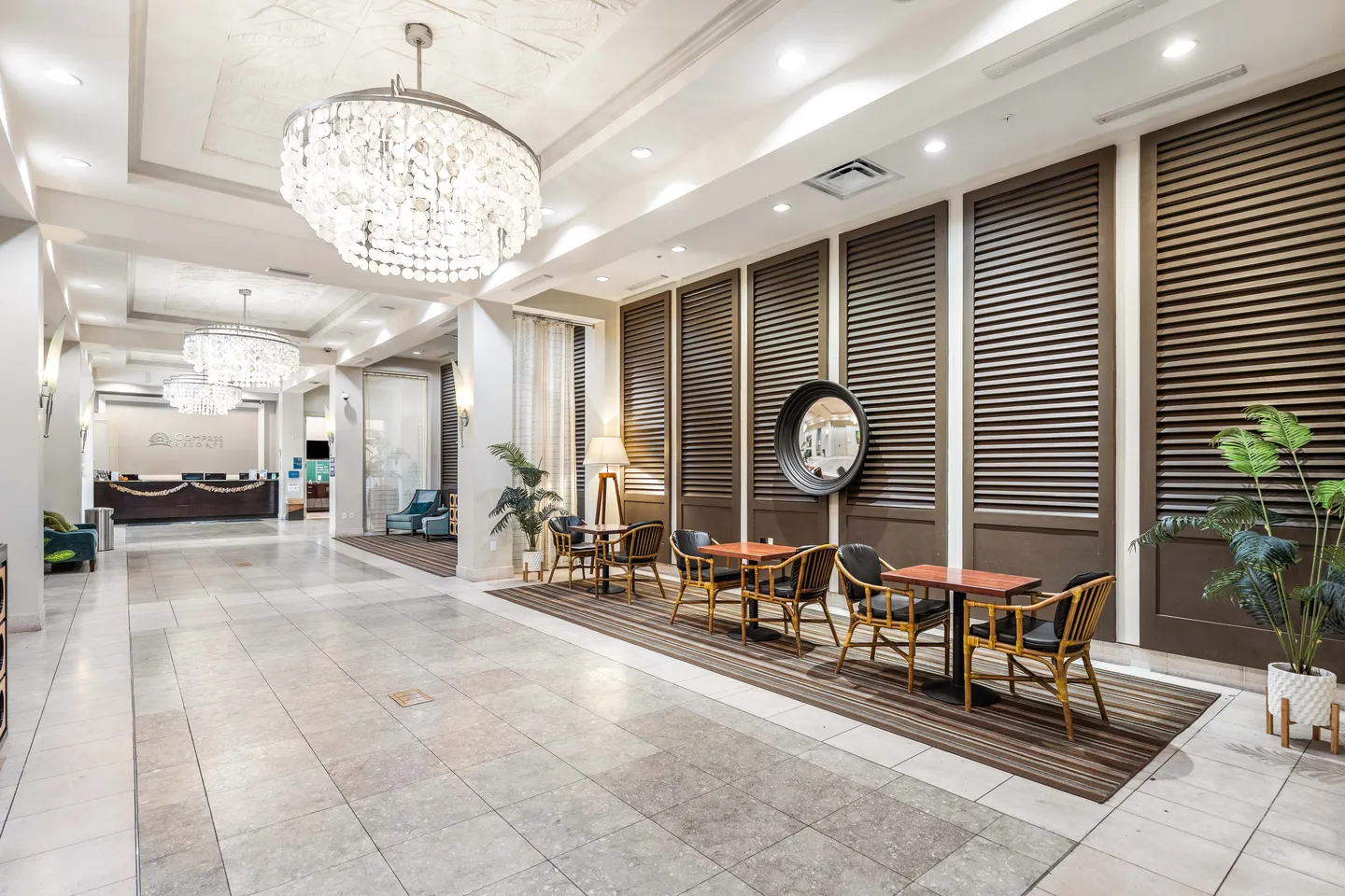 Spacious building lobby featuring tiered crystal chandeliers, tile flooring, and small dining or seating tables along a wall with slatted panels.