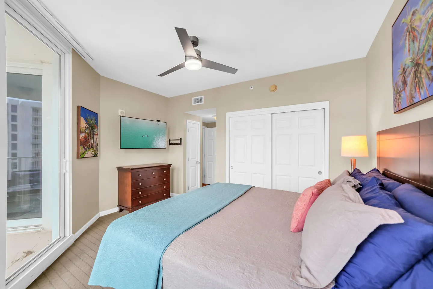Bedroom featuring a made bed, wall-mounted television over a wooden dresser, sliding closet doors, and a glass door opening to a balcony.