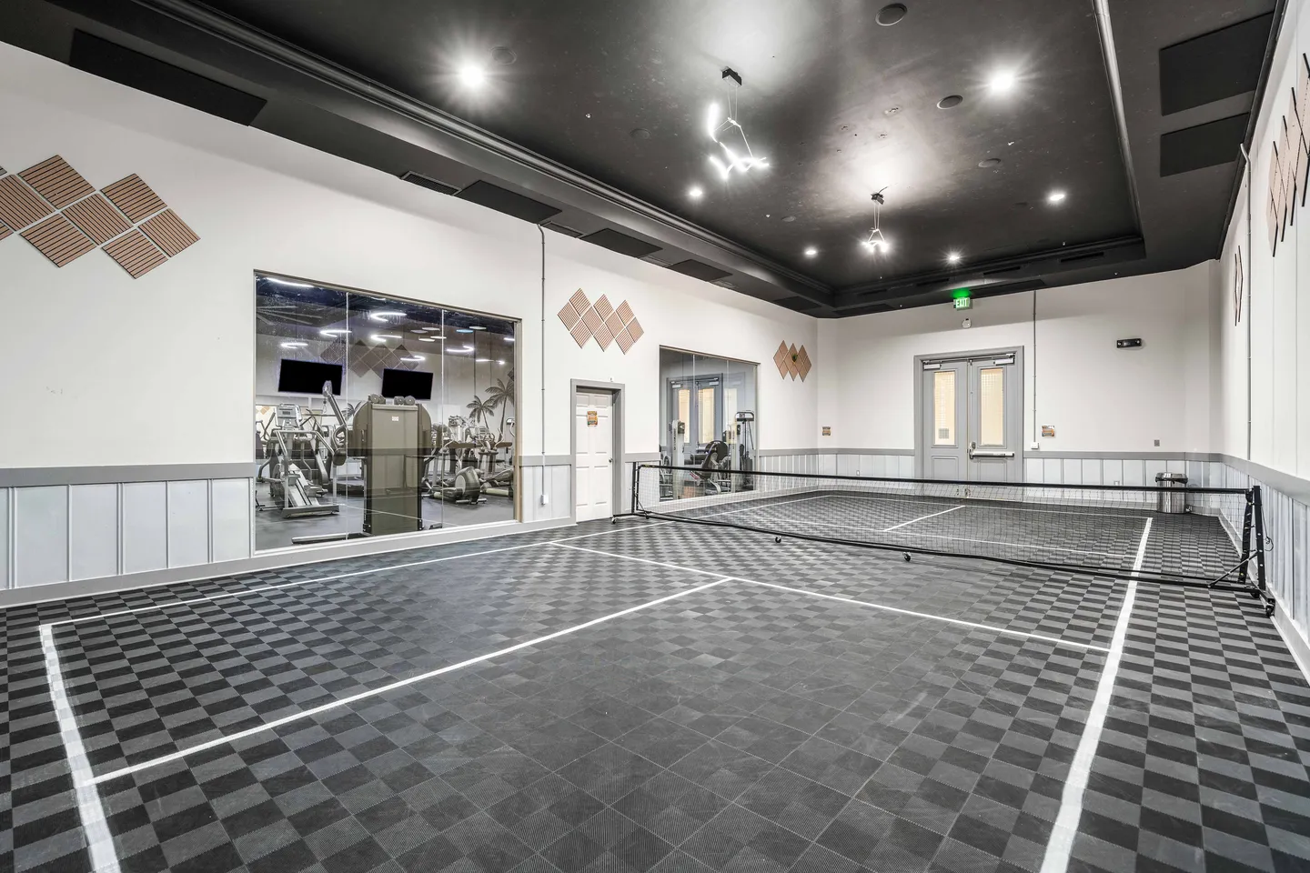 Indoor sports court with checkered flooring and a net, featuring large windows showing an adjacent fitness center.