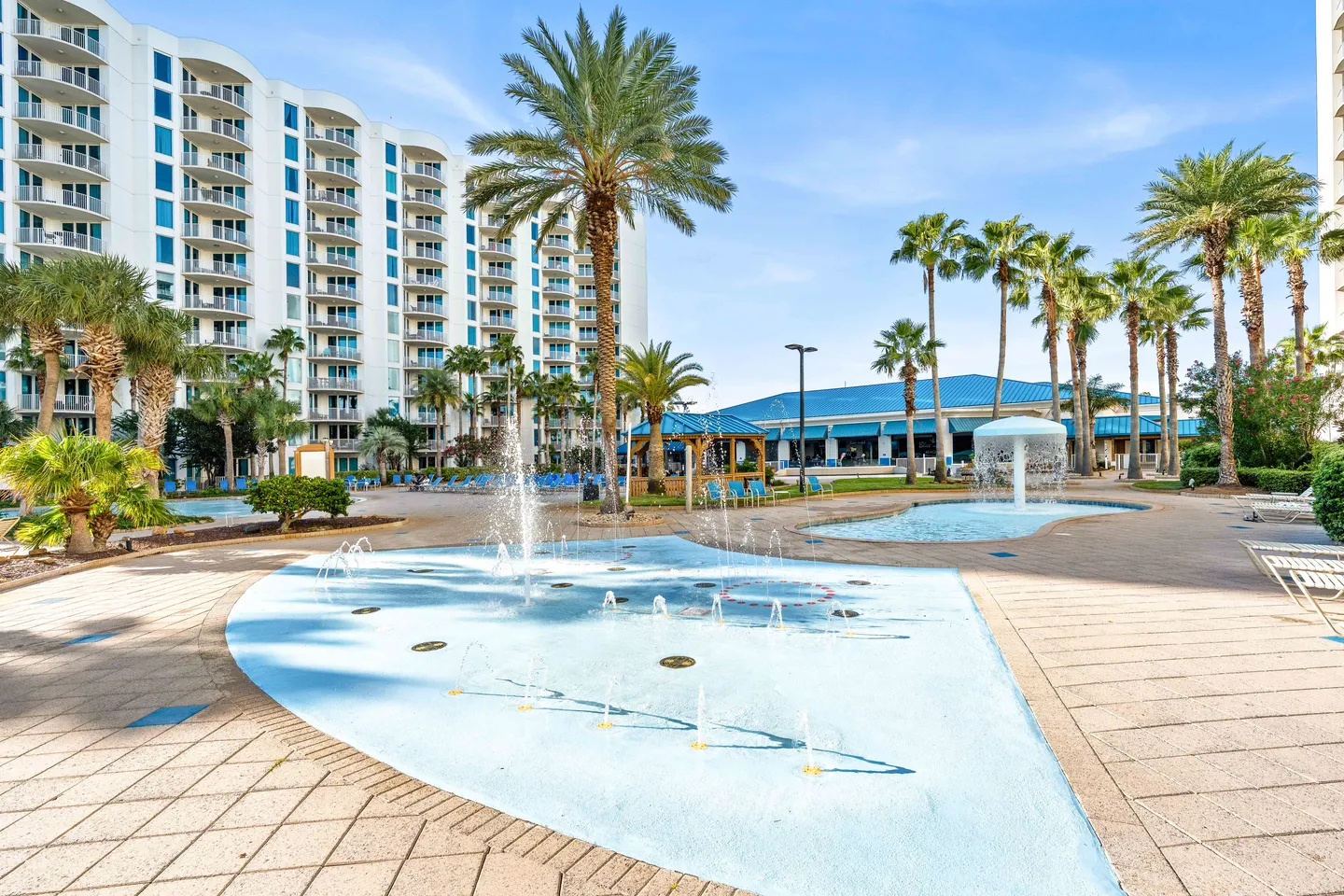 Outdoor resort splash pad with water fountains in front of palm trees and a multi-story building under a blue sky.