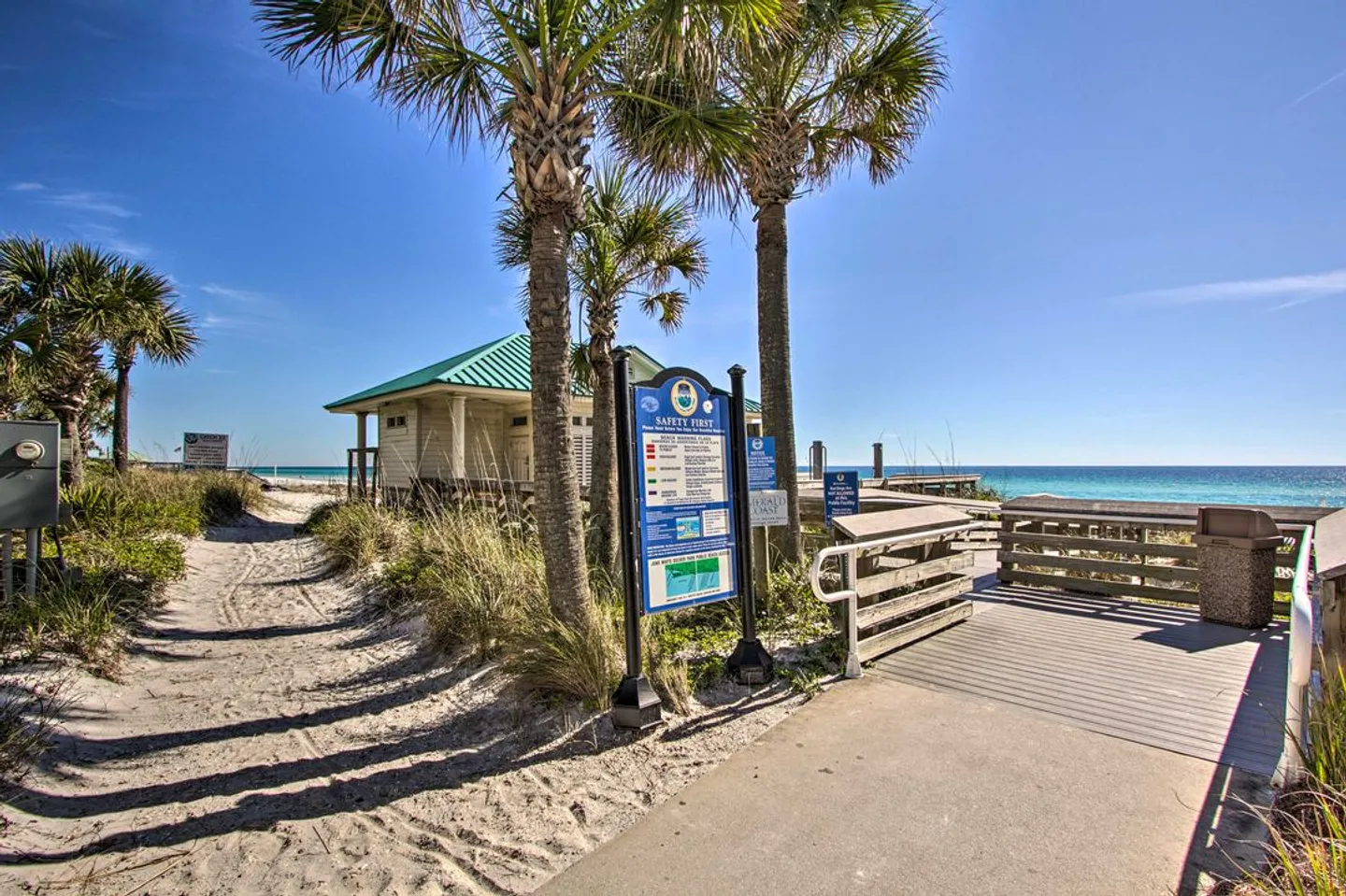 Boardwalk and sandy path leading to the beach past palm trees, informational signs, and a small pavilion.
