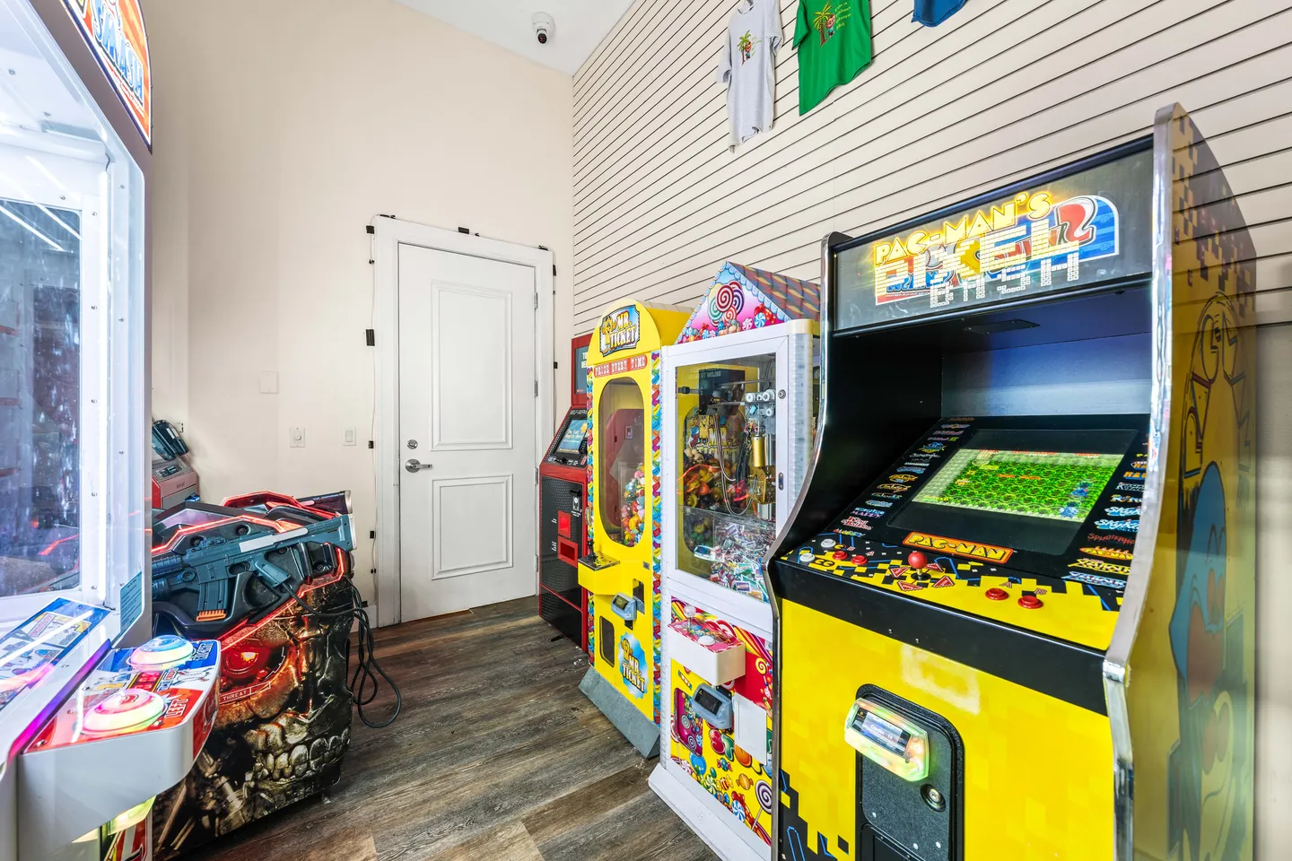 Game room with several arcade machines and claw games lined up against slatwall and plain walls.