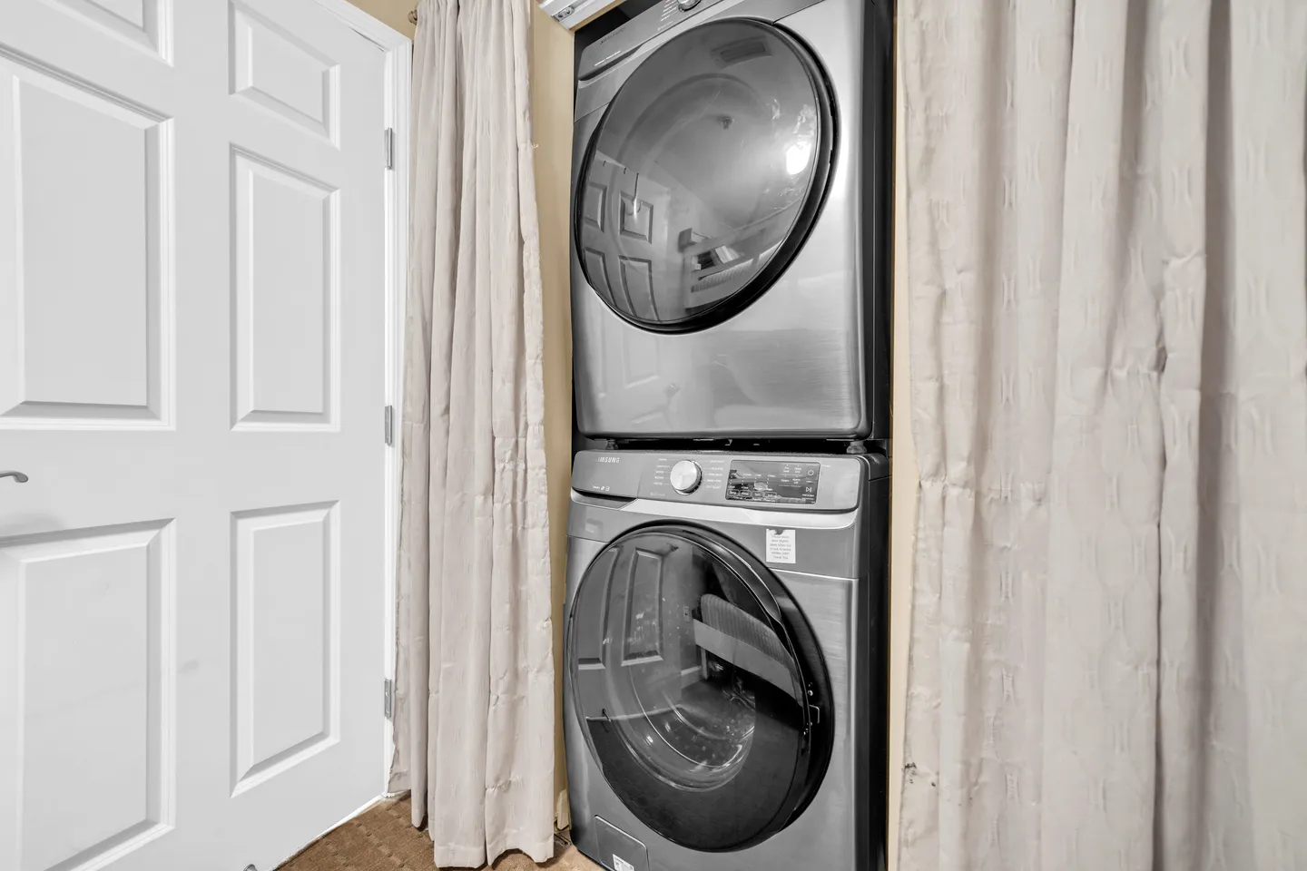 Stackable metallic front-load washer and dryer set framed by light curtains next to a white door.