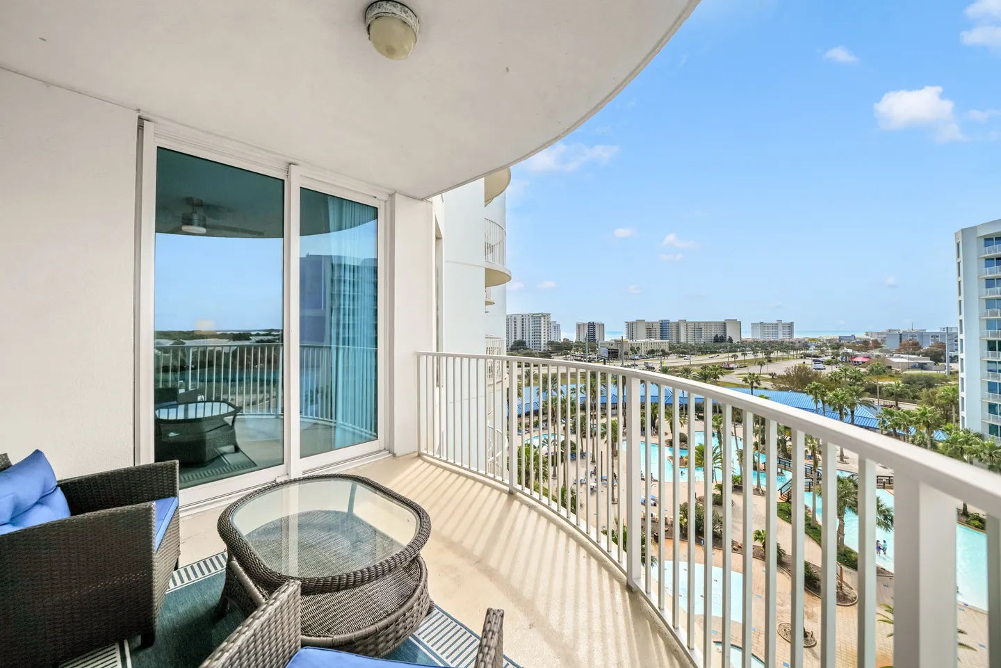 Curved high-rise balcony with outdoor wicker seating, overlooking resort swimming pools and the beach.