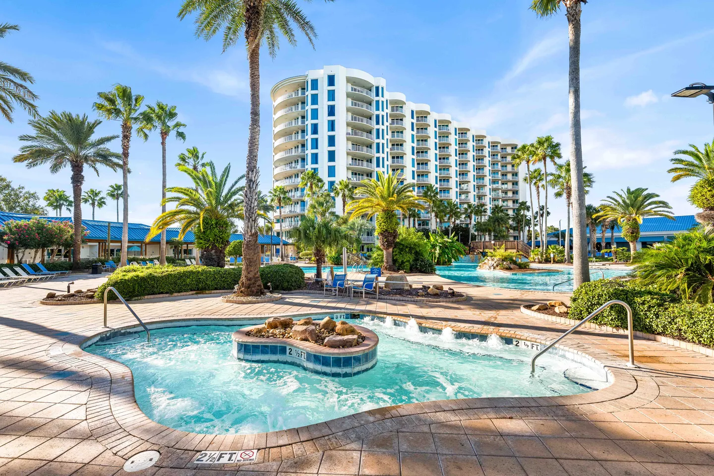 Outdoor resort pool and hot tub area surrounded by palm trees with a tall multi-story building in the background.