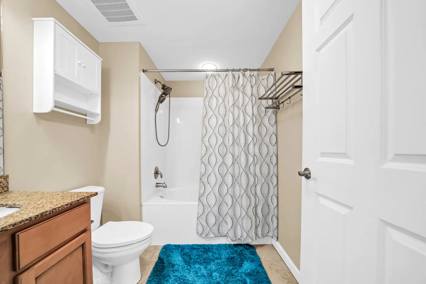 Bathroom with a vanity, toilet, shower and tub combination with a patterned curtain, and a blue rug on tiled flooring.