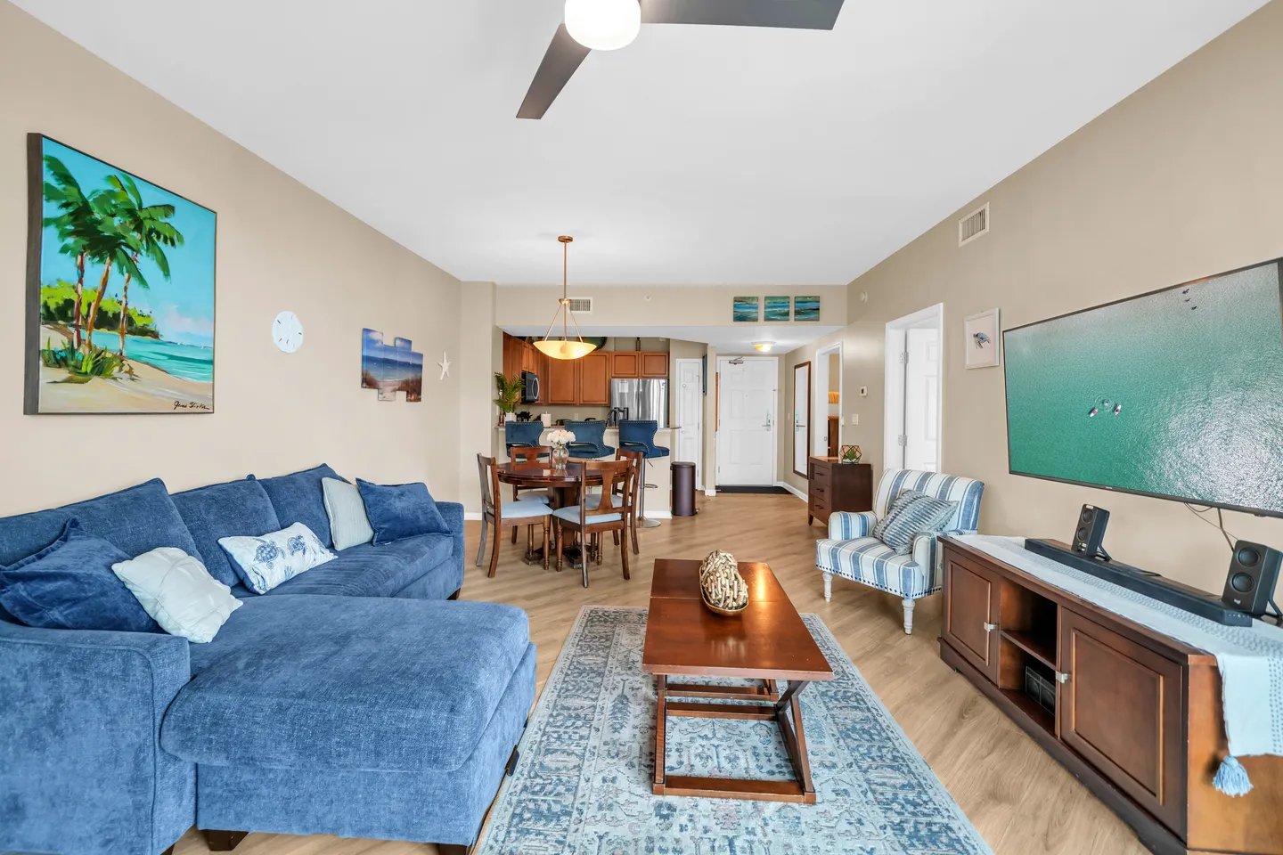 Open-concept living room with a blue sectional sofa, coffee table, TV console, dining area, and kitchen in the background.