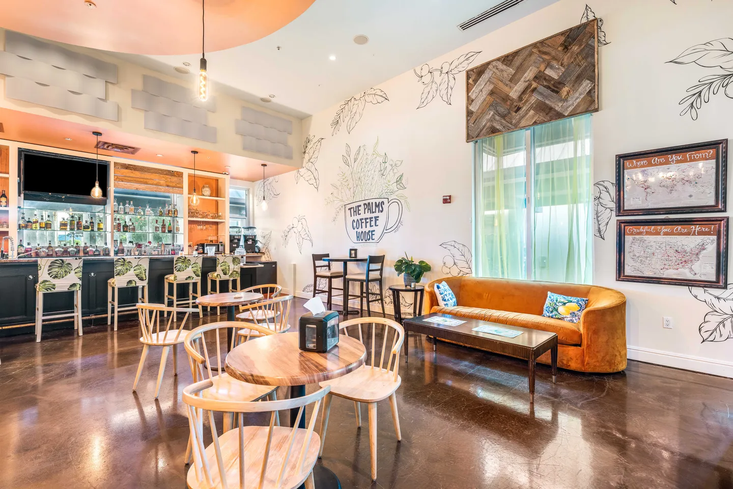 Indoor lounge and cafe area featuring small round tables with chairs, an orange sofa, wall murals, and a bar counter with barstools.