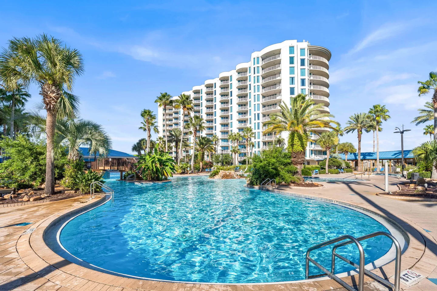 Outdoor resort-style swimming pool surrounded by palm trees in front of a multi-story white condominium tower under a clear blue sky.
