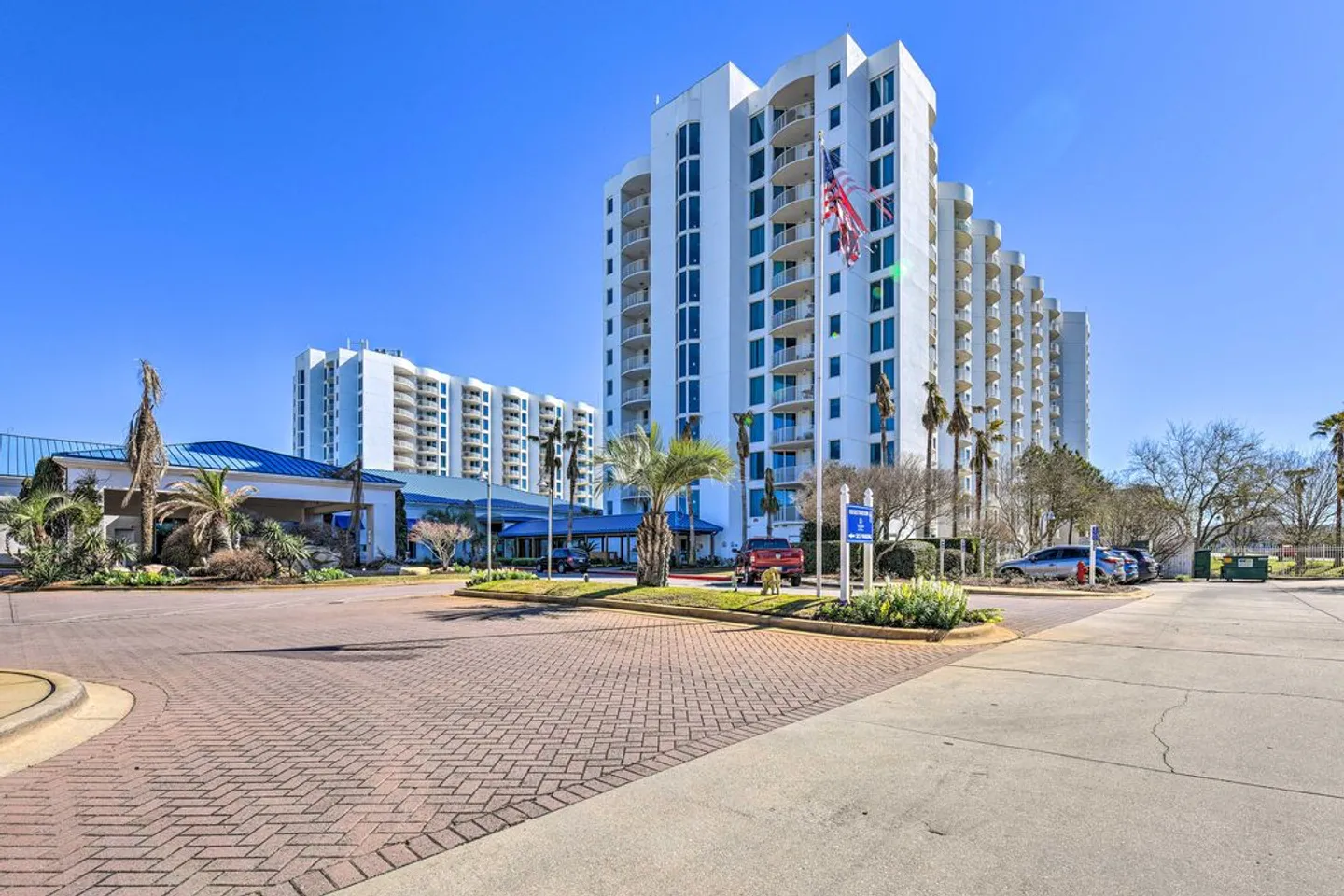 Exterior view of a multi-story white condominium complex with a paved driveway and palm trees under a clear blue sky.