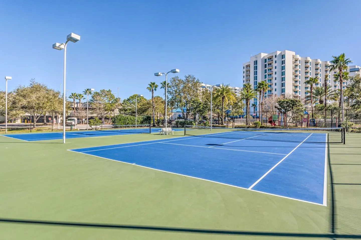 Outdoor tennis and pickleball courts with blue and green surfaces under a clear blue sky, surrounded by fencing and trees with multi-story buildings in the background.
