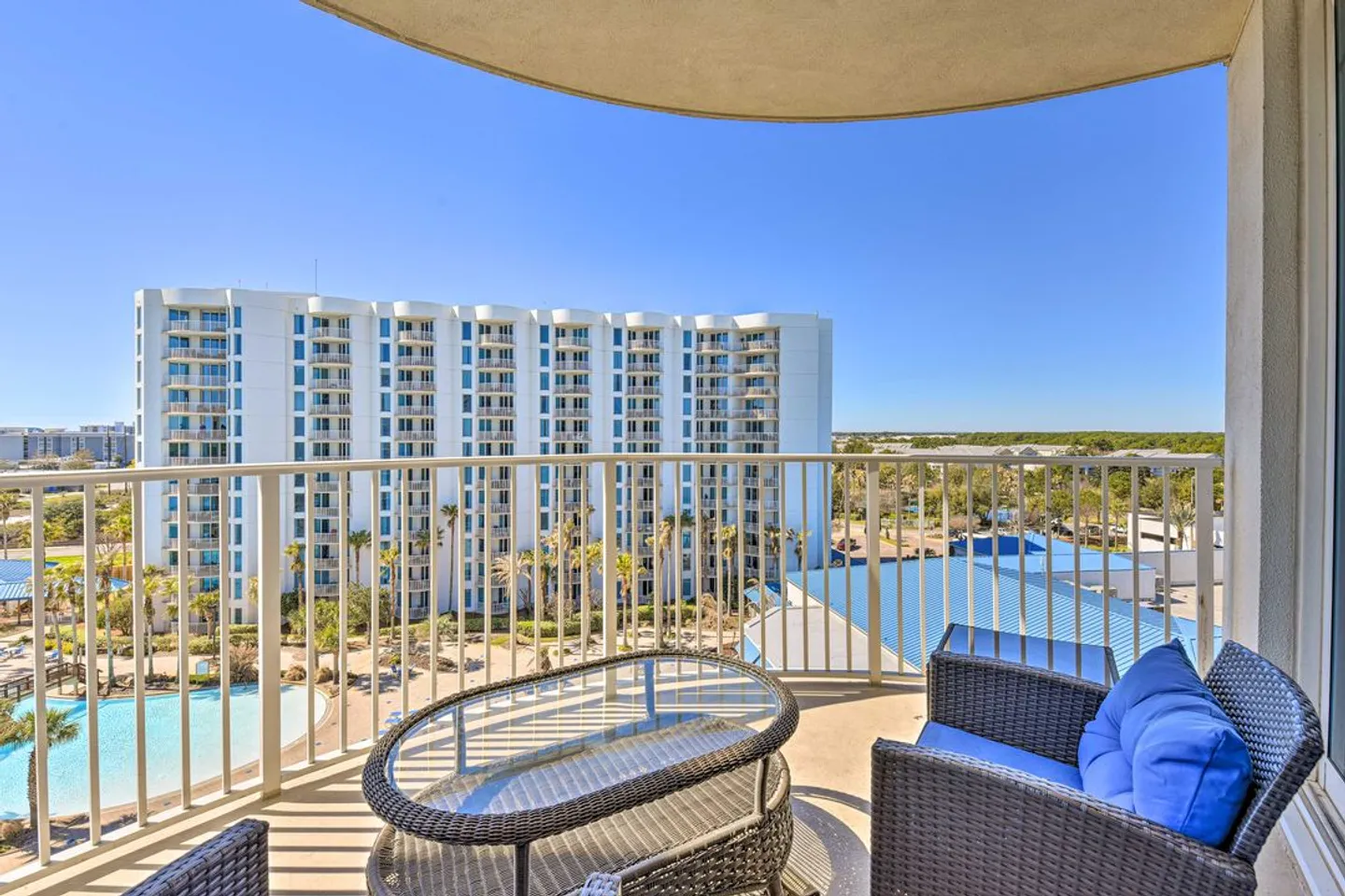 Balcony view overlooking a swimming pool area and a multi-story resort building under a clear blue sky.