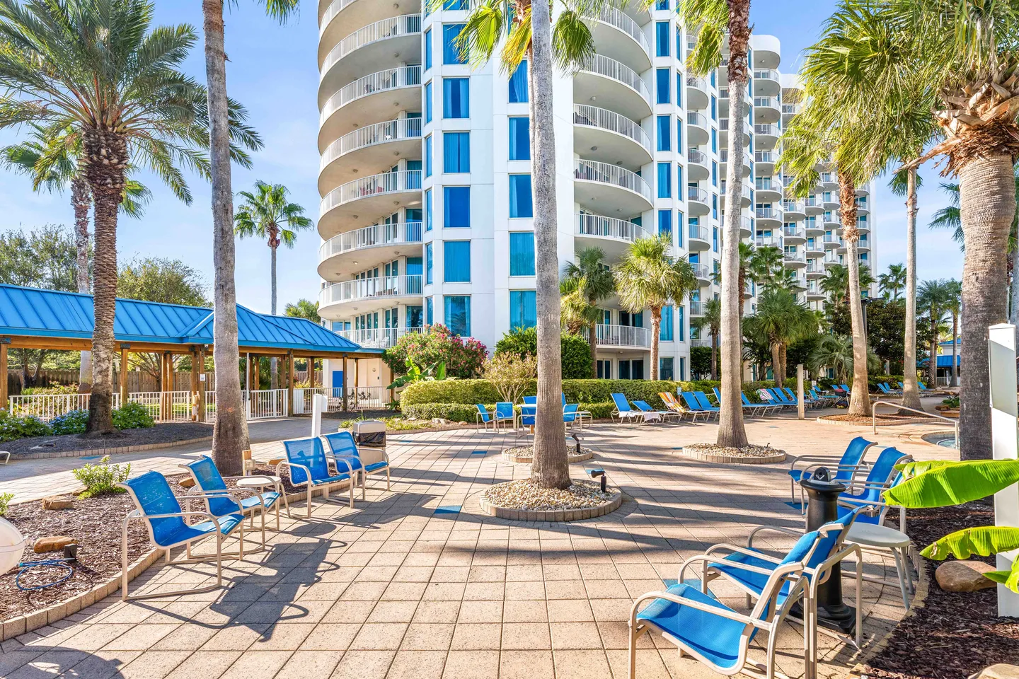 Paved outdoor patio area with blue armchairs, tall palm trees, and a high-rise condominium building in the background.