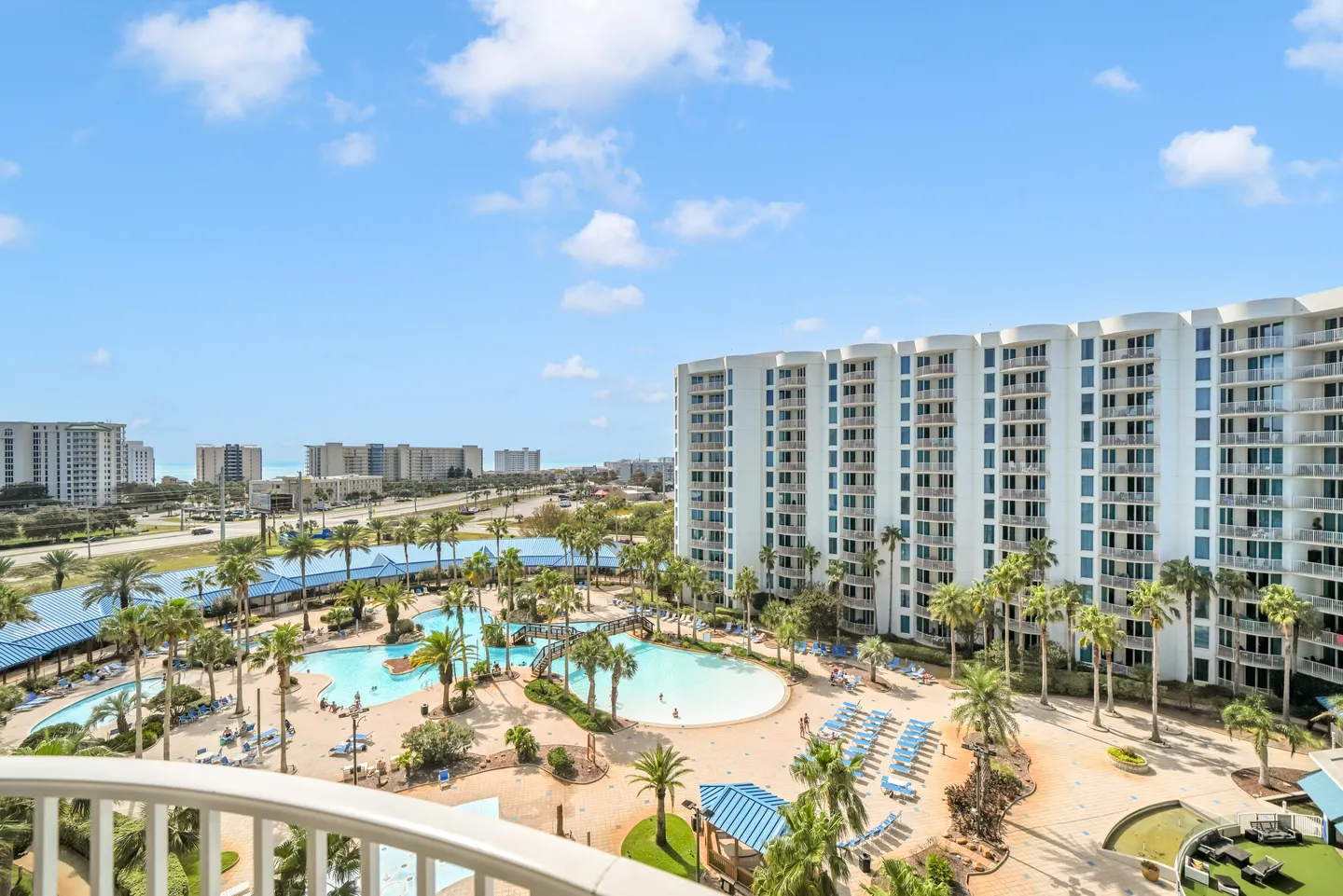 Balcony view overlooking a resort-style outdoor pool complex, palm trees, and multi-story condominium buildings under a sunny blue sky.