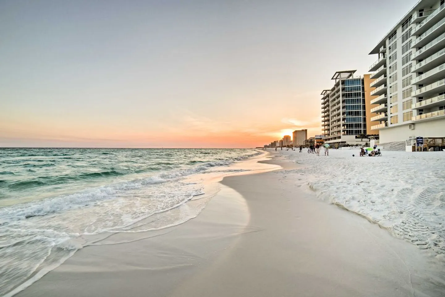 A wide view of a white sandy beach and ocean surf beside high-rise beachfront condominium buildings at sunset.