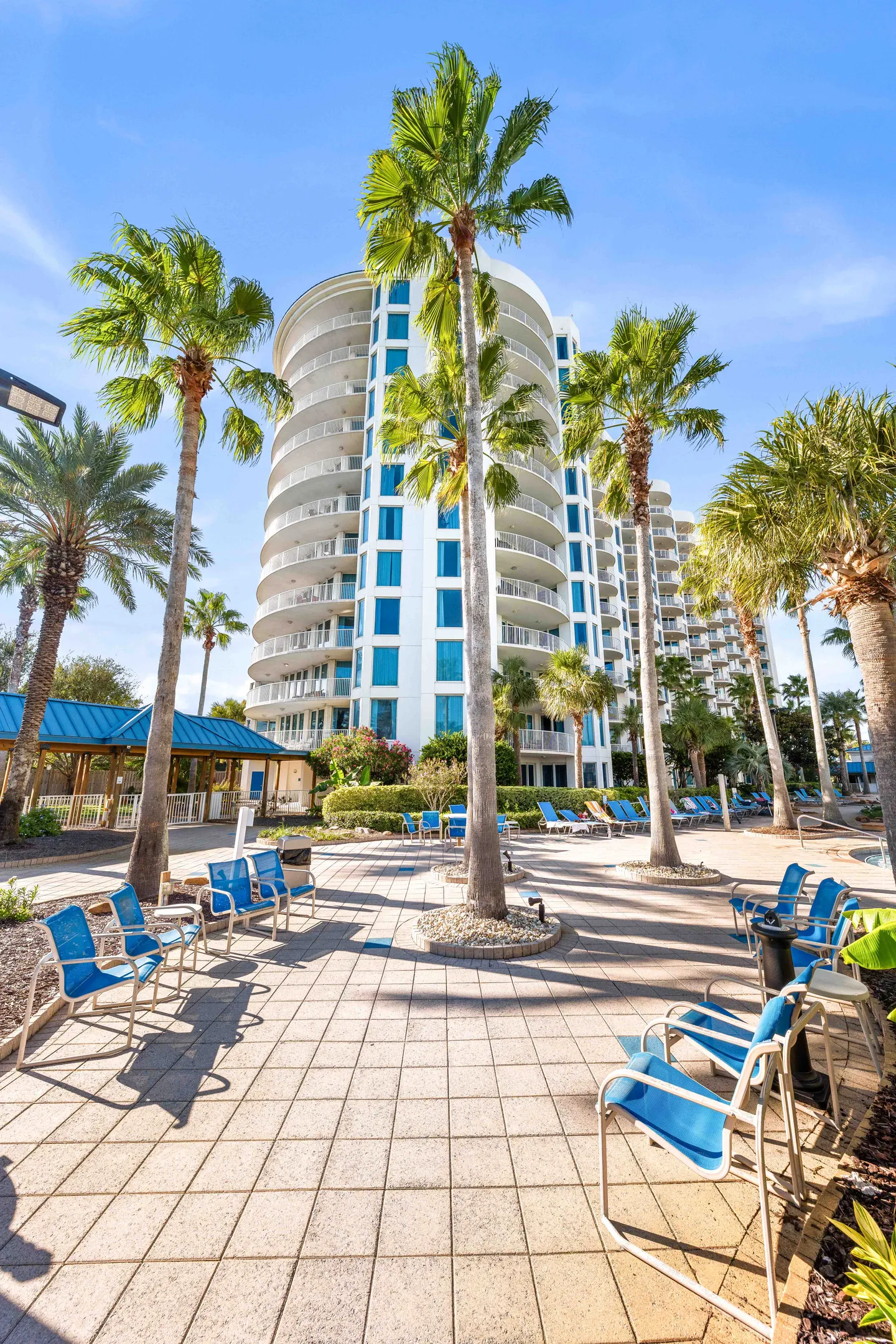 Outdoor paved patio area with blue chairs and tall palm trees in front of a modern high-rise resort building under a clear blue sky.