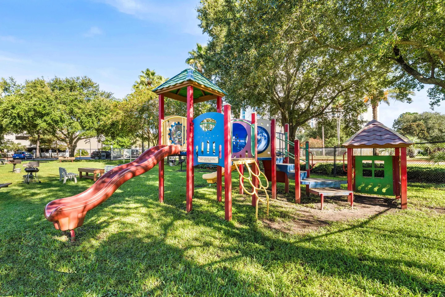 Outdoor children's playground structure with a red slide on a grassy lawn surrounded by trees.