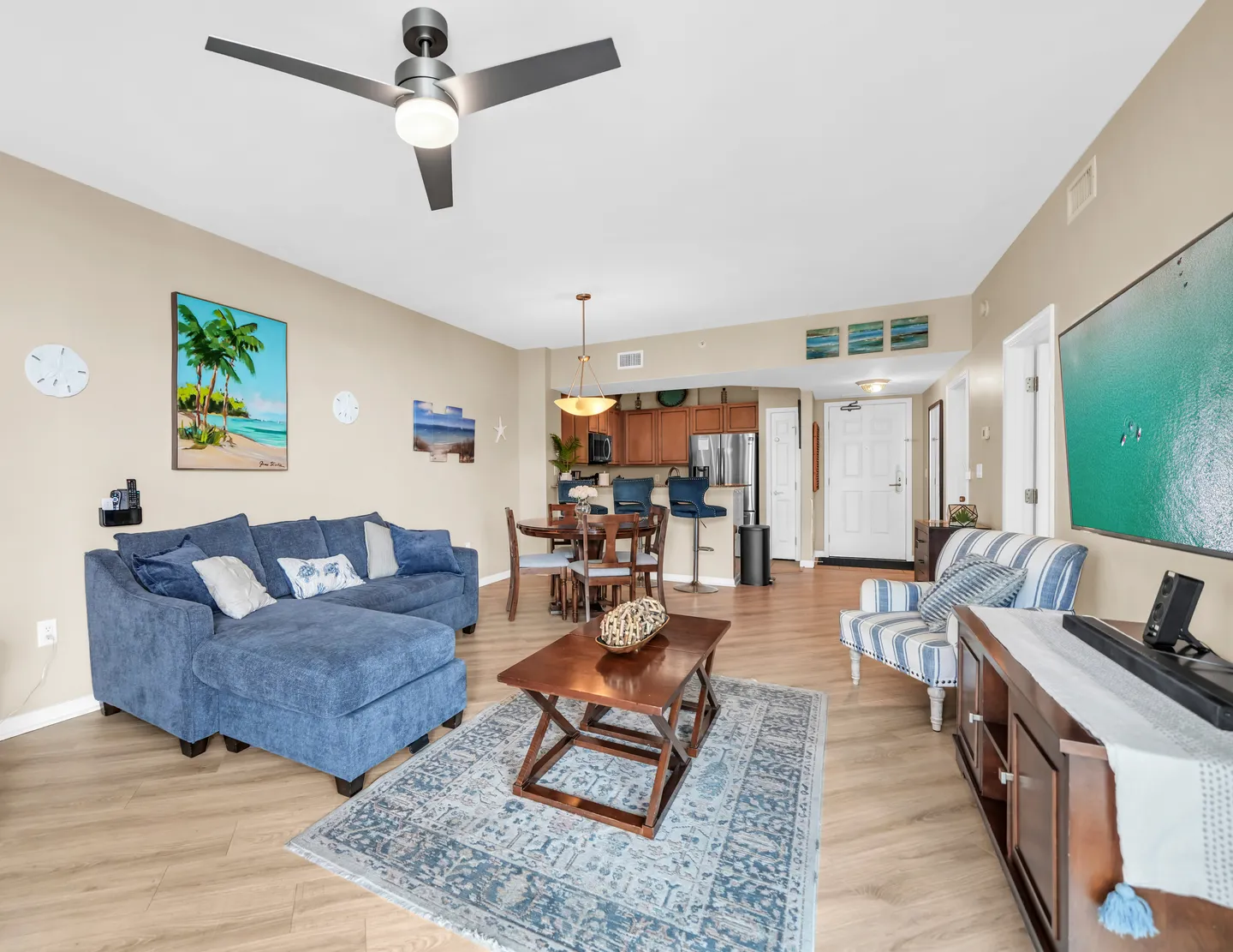 Open-concept living room with a blue sectional sofa, patterned area rug, coffee table, striped accent chair, dining area, and kitchen in the background.