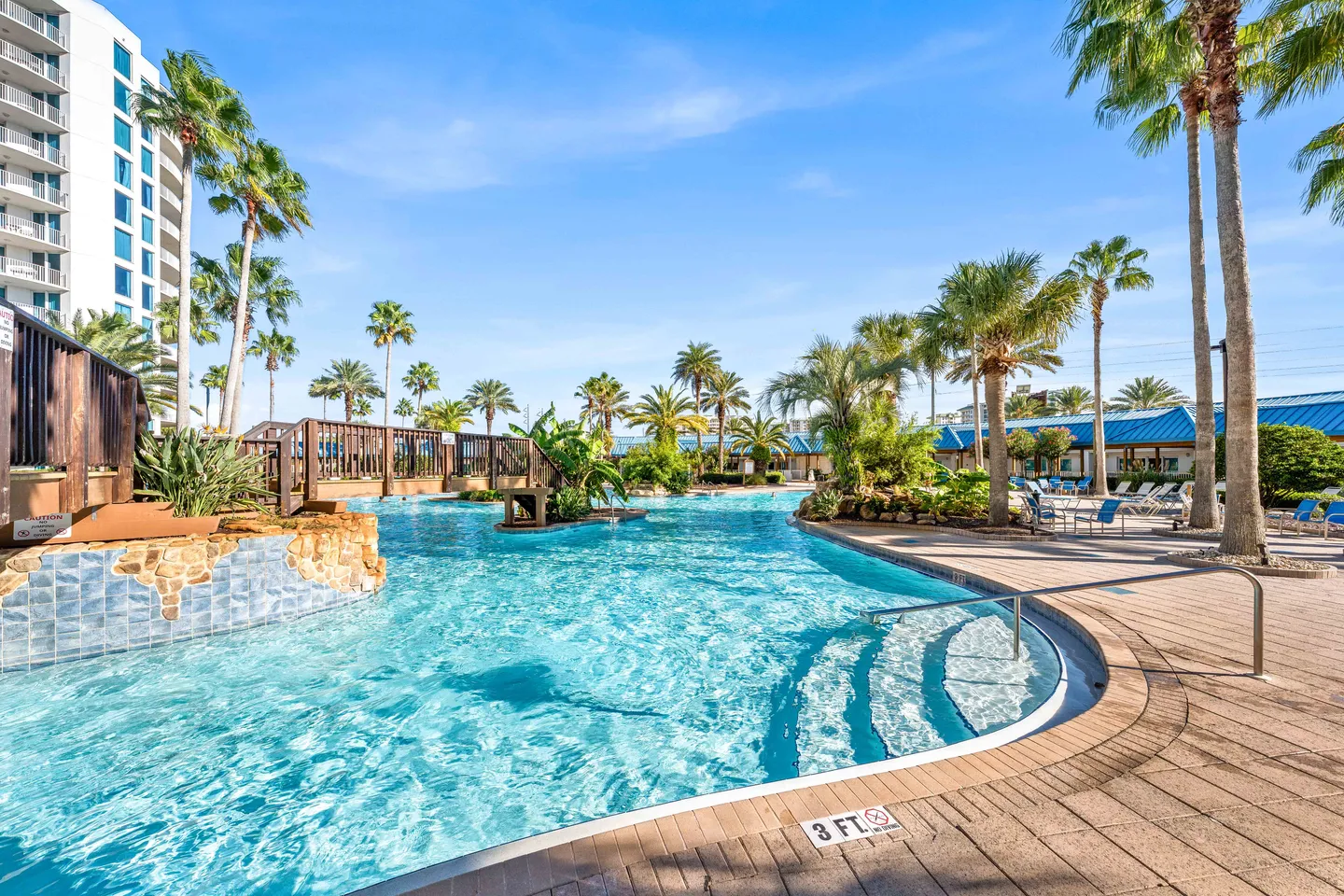 Large outdoor resort swimming pool with a pedestrian footbridge, curved entry steps, and surrounding palm trees under a blue sky.