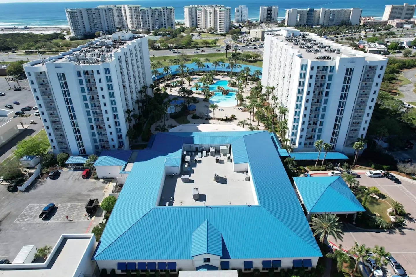 Aerial view of a coastal resort complex featuring white multistory towers, a bright blue metal roof, and an expansive courtyard with outdoor swimming pools.