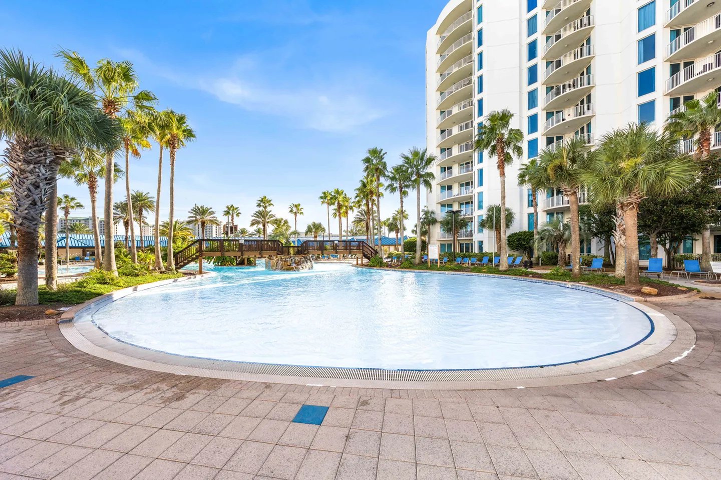 Large zero-entry outdoor resort pool with a wooden pedestrian bridge, palm trees, and a multistory white condominium building.