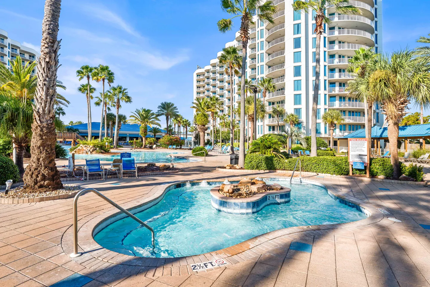 Outdoor hot tub and resort pool complex surrounded by palm trees and multi-story condominium buildings under a blue sky.