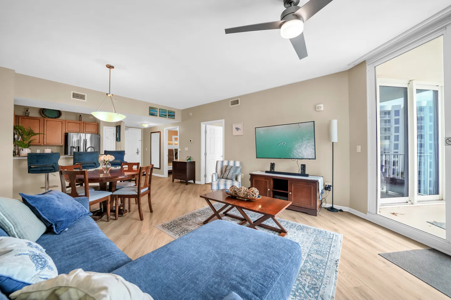 Open-concept living room with a blue sectional sofa, a wooden coffee table, a mounted flat-screen TV, a dining table, and a kitchen bar.