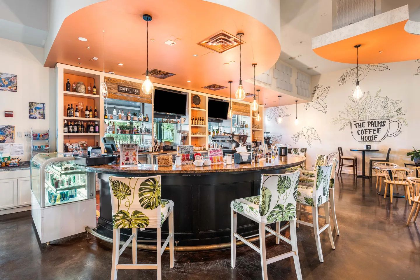 A curved bar counter with tropical-patterned barstools in a coffee shop labeled The Palms Coffee House.