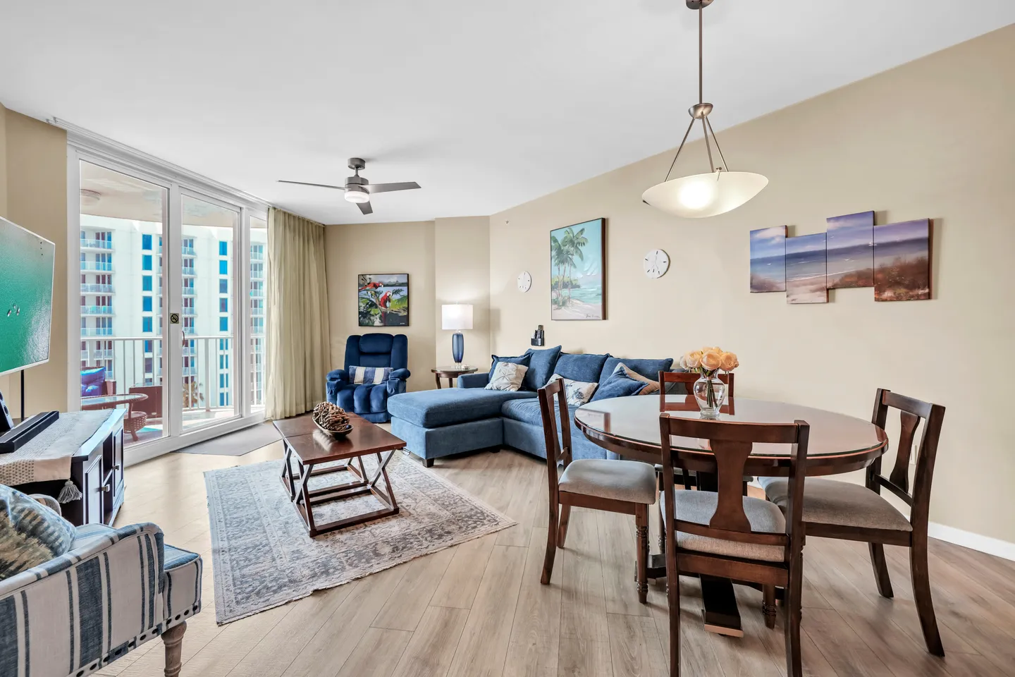 Open living and dining area with wood-look flooring, a blue sectional couch, dining table with chairs, and sliding glass doors to a balcony.