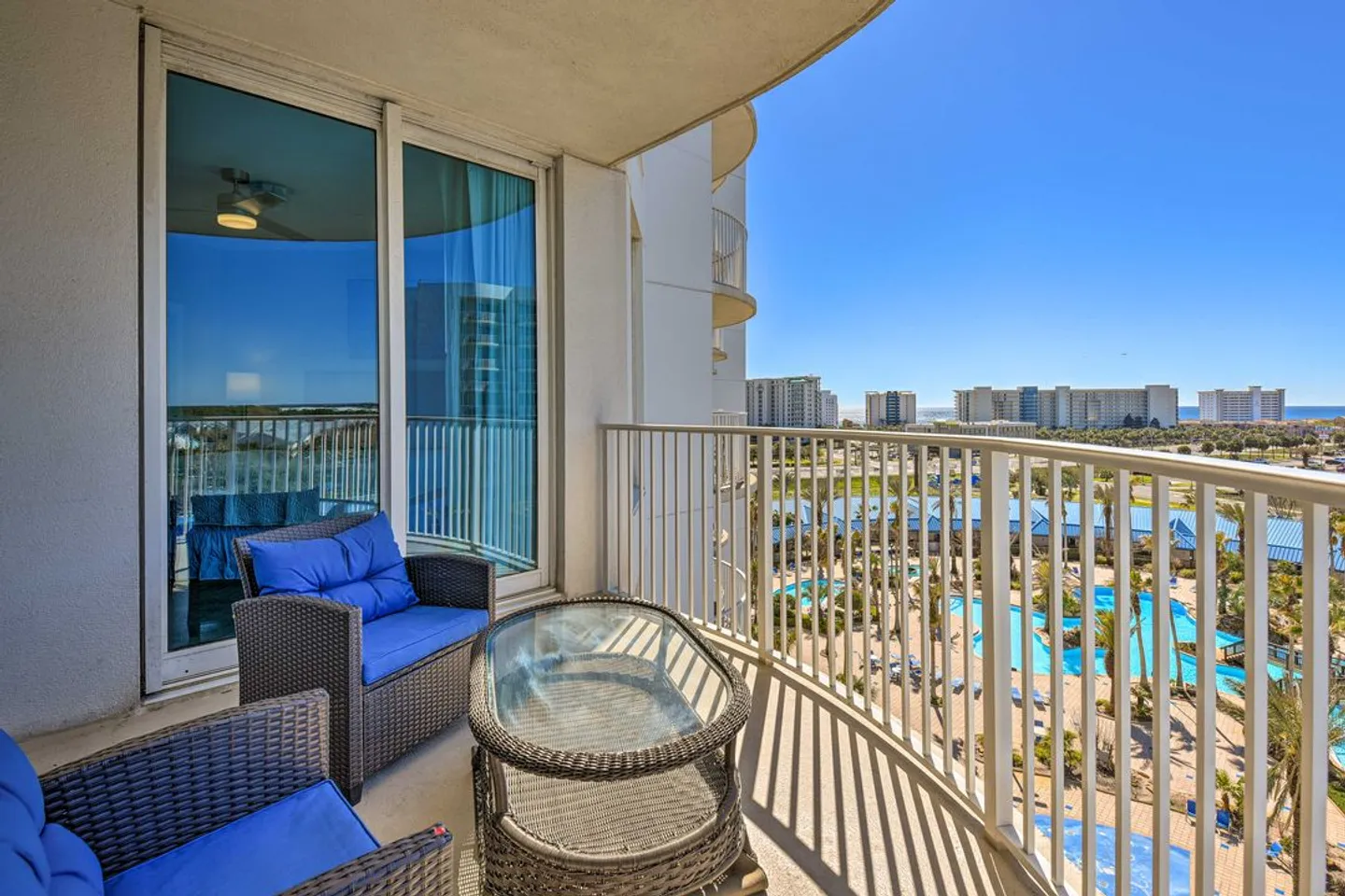 Balcony with wicker armchairs, glass-top table, and railing overlooking a resort pool area and the ocean under a clear blue sky.