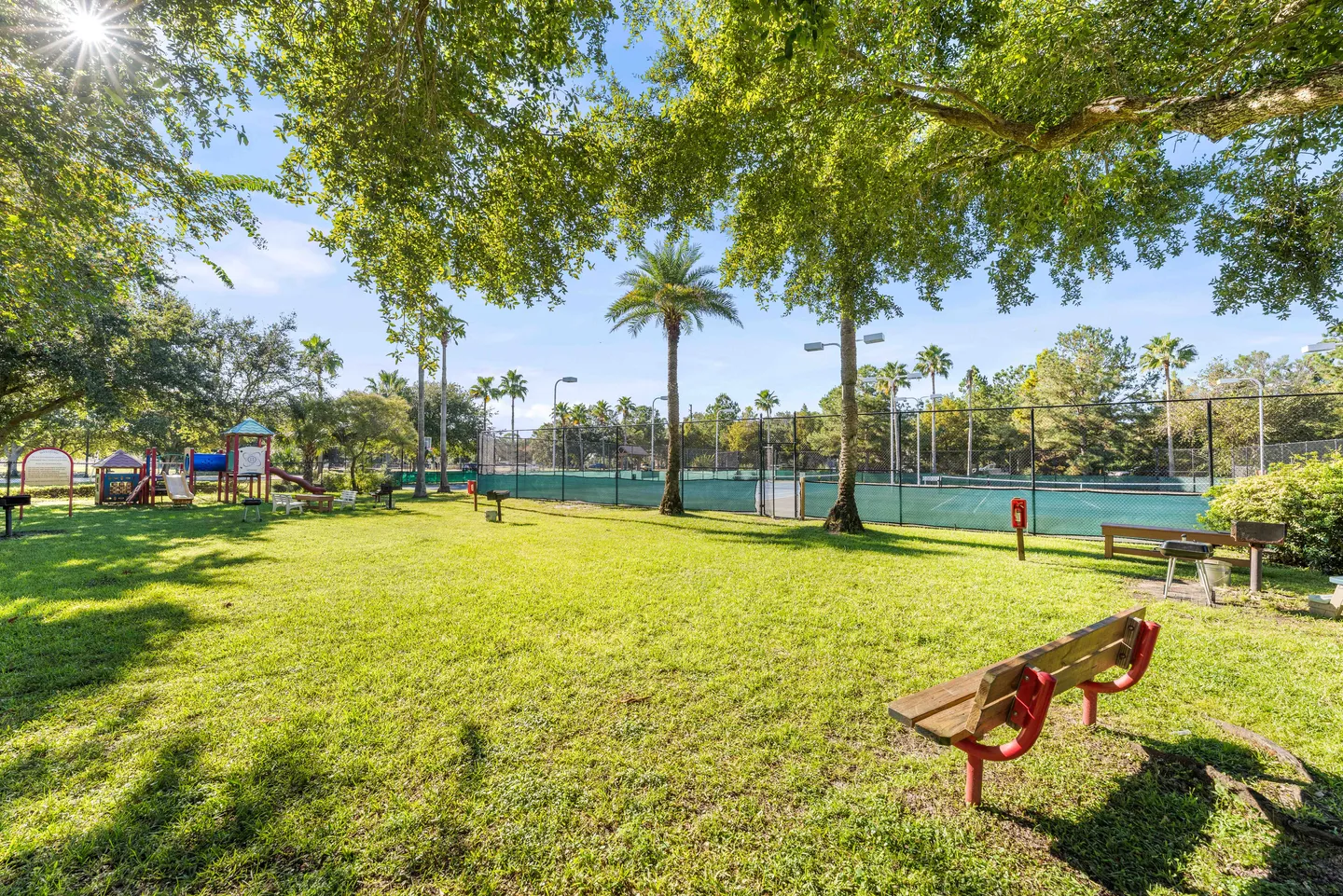 Outdoor community park area with a grassy lawn, park bench, playground set, and fenced tennis courts under a sunny sky.