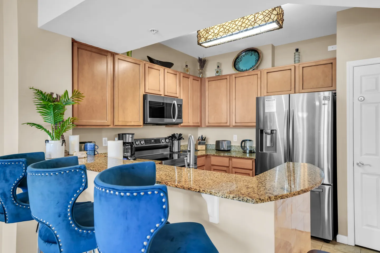 Kitchen with light wood cabinetry, speckled countertops, stainless steel appliances, and a breakfast bar with tufted blue velvet stools.