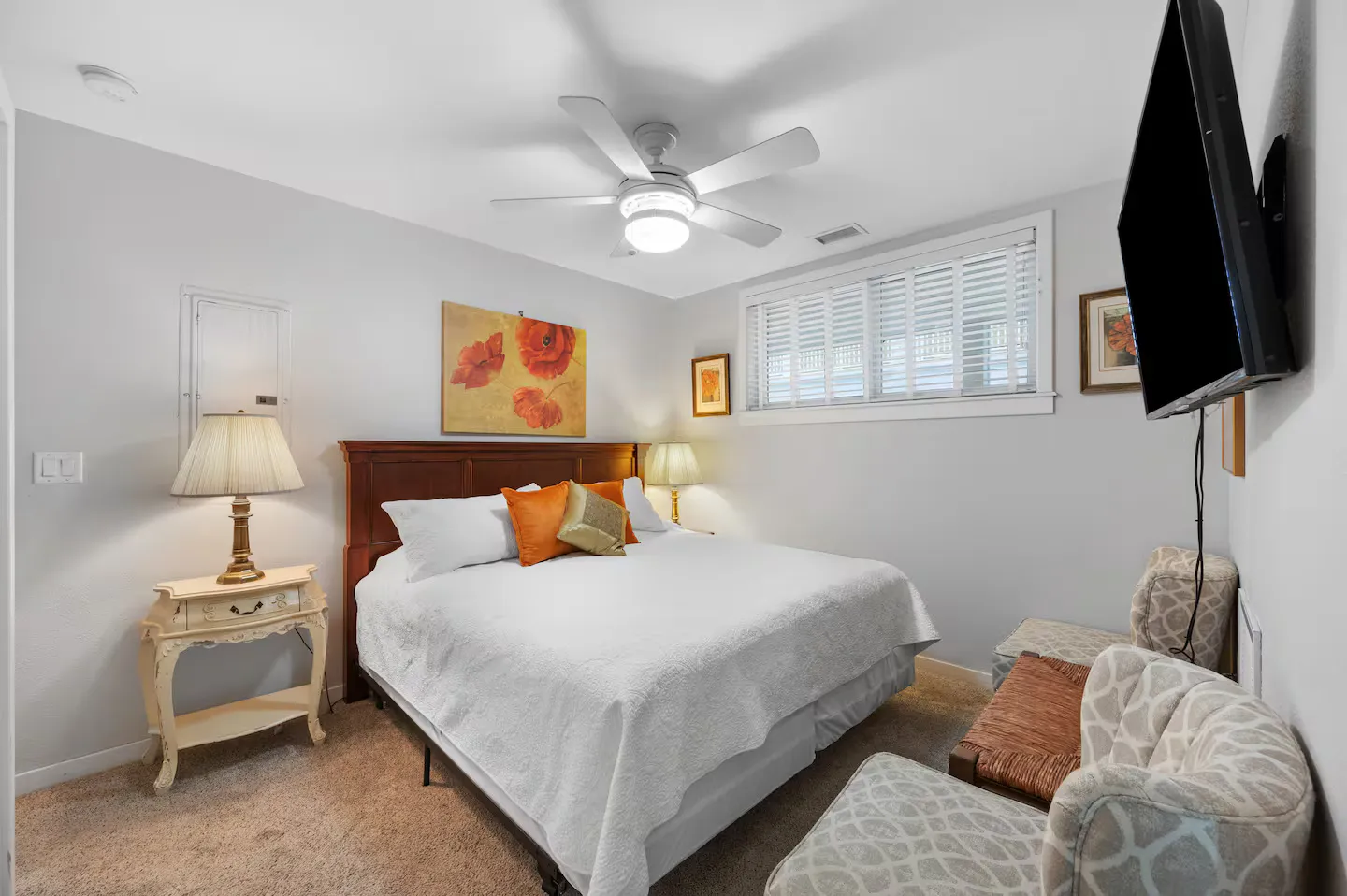 Bedroom featuring a made bed with a dark wood headboard, floral wall art, side tables with lamps, a ceiling fan, and armchairs.