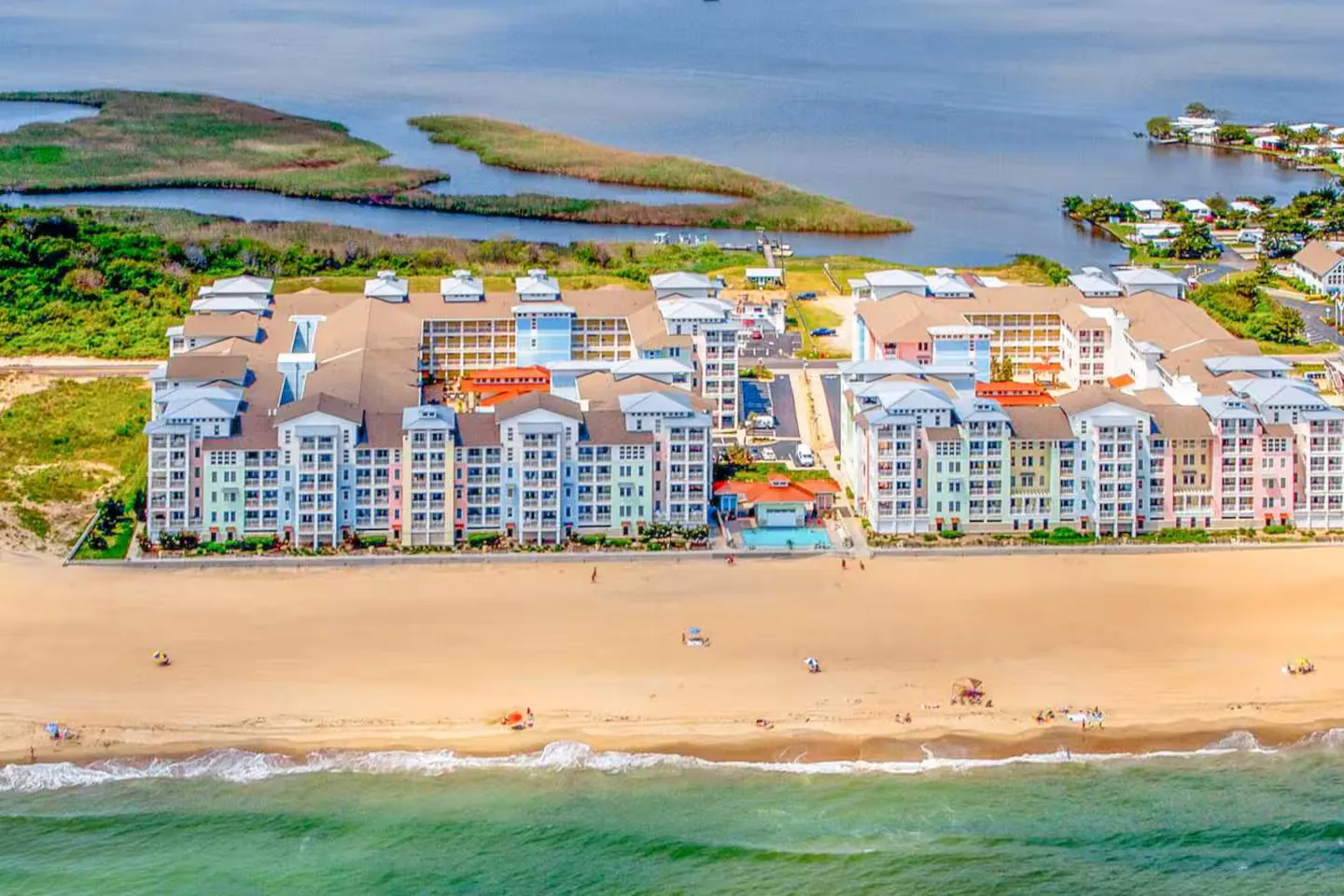 Aerial view of a beachfront condominium resort complex facing a sandy beach and ocean, with a bay behind it.