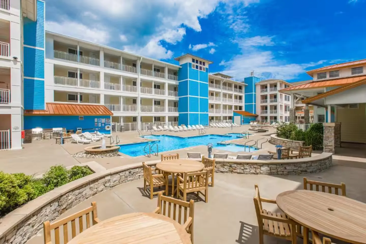 Outdoor swimming pool and patio dining area framed by multi-story blue and white resort buildings under a blue sky.