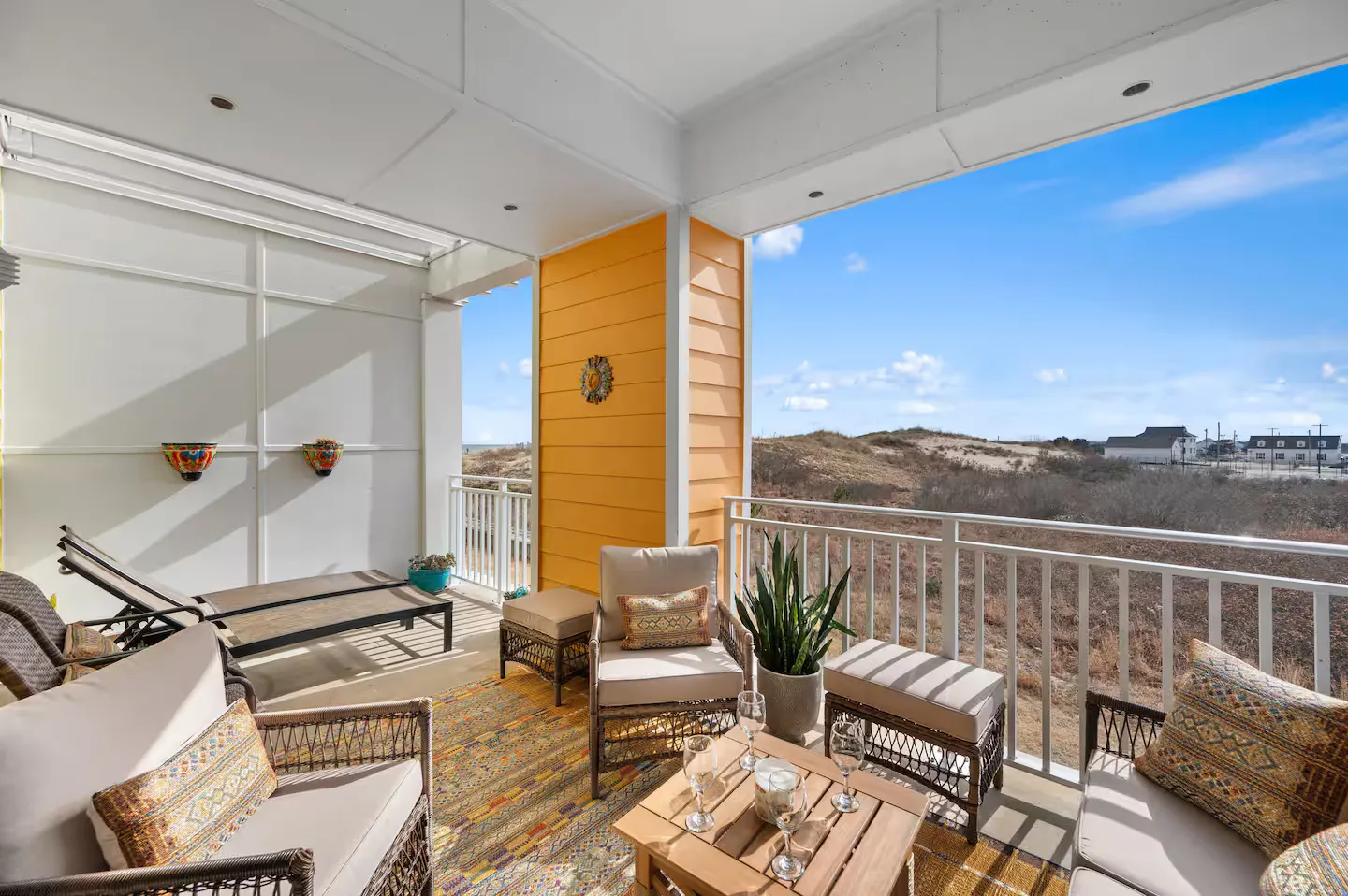 Covered outdoor patio with wicker seating, lounge chairs, a coffee table, and views of coastal dunes under a blue sky.