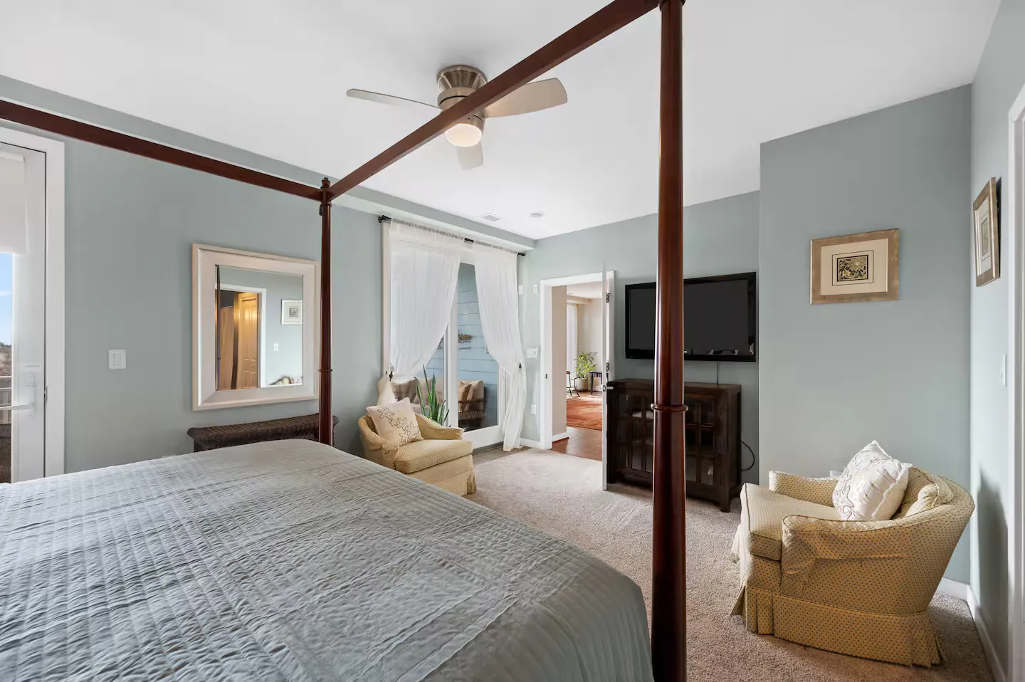 Bedroom featuring a four-poster canopy bed, light blue walls, carpeted floor, armchairs, and a wall-mounted TV.