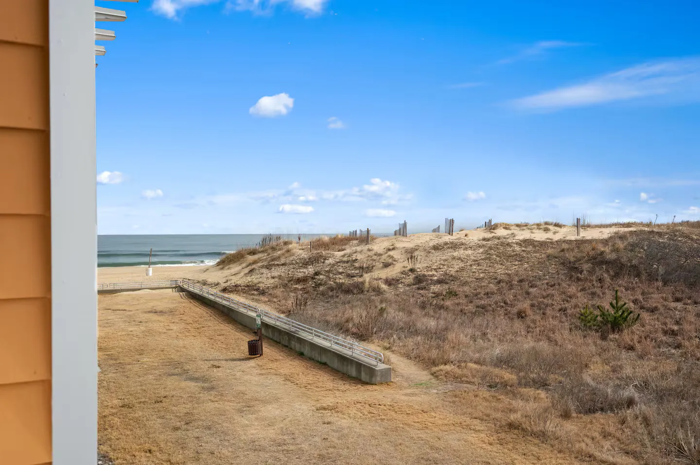 View of sand dunes, a walkway, and the ocean under a blue sky, framed by an exterior building wall.