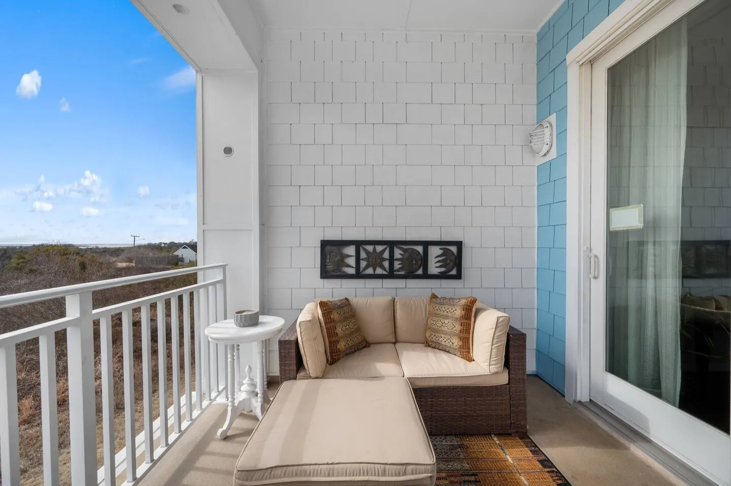 A covered balcony with an outdoor sectional sofa, an ottoman, a small white side table, and railings overlooking distant views.
