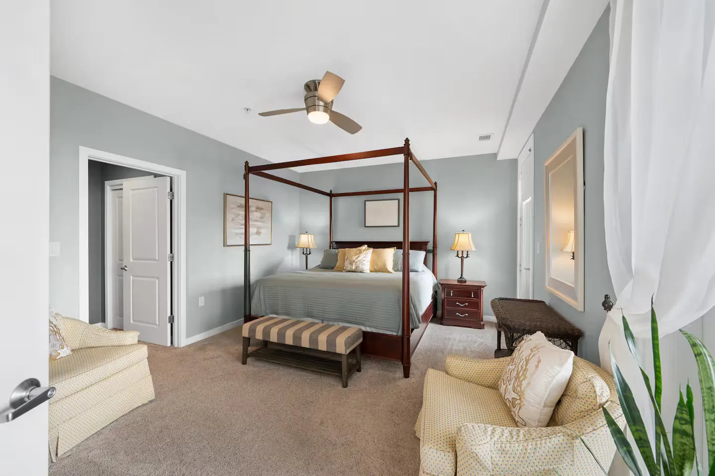 Bedroom featuring a dark wood canopy bed, soft blue walls, carpeted floor, ceiling fan, and two upholstered armchairs.