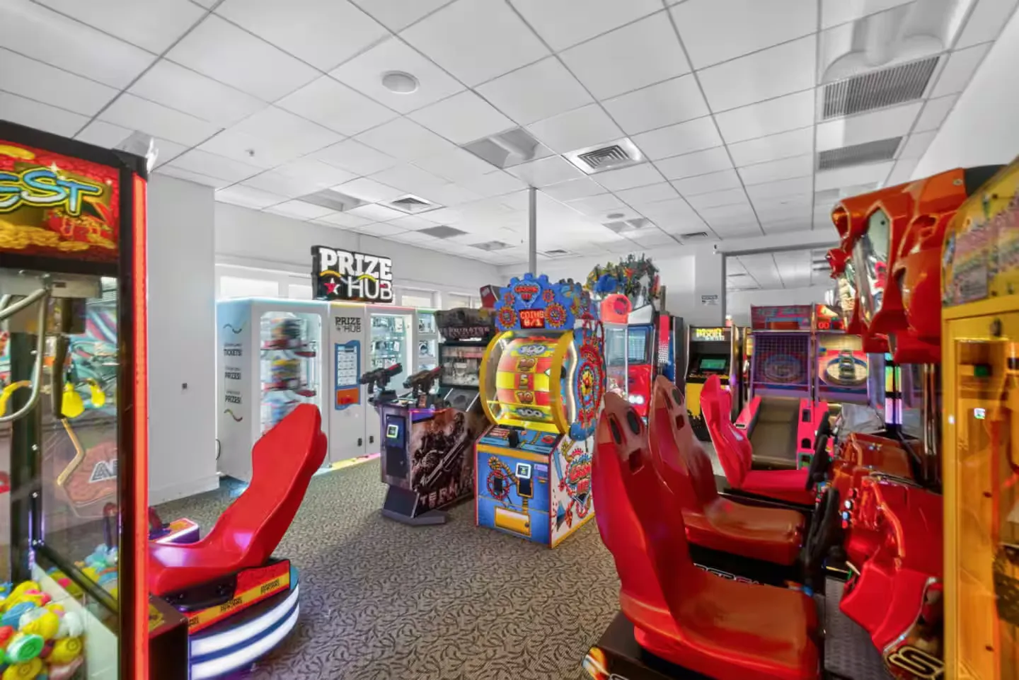 Indoor arcade room filled with various electronic games, prize machines, and racing simulators with red bucket seats.