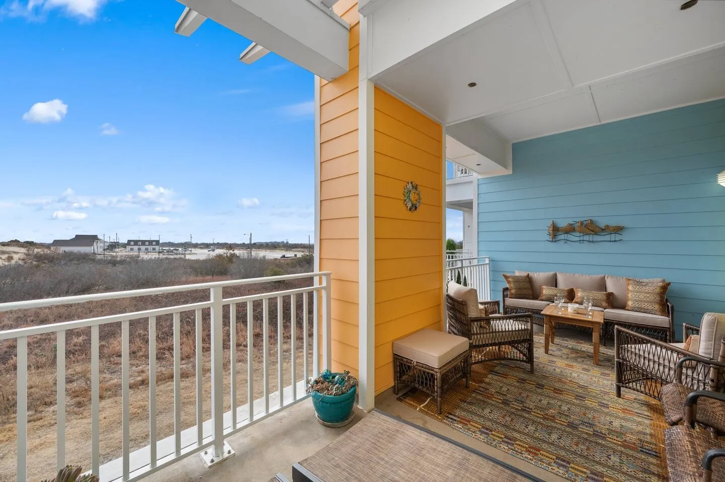Covered outdoor patio with a sofa, armchairs, coffee table, and area rug overlooking open land under a blue sky.