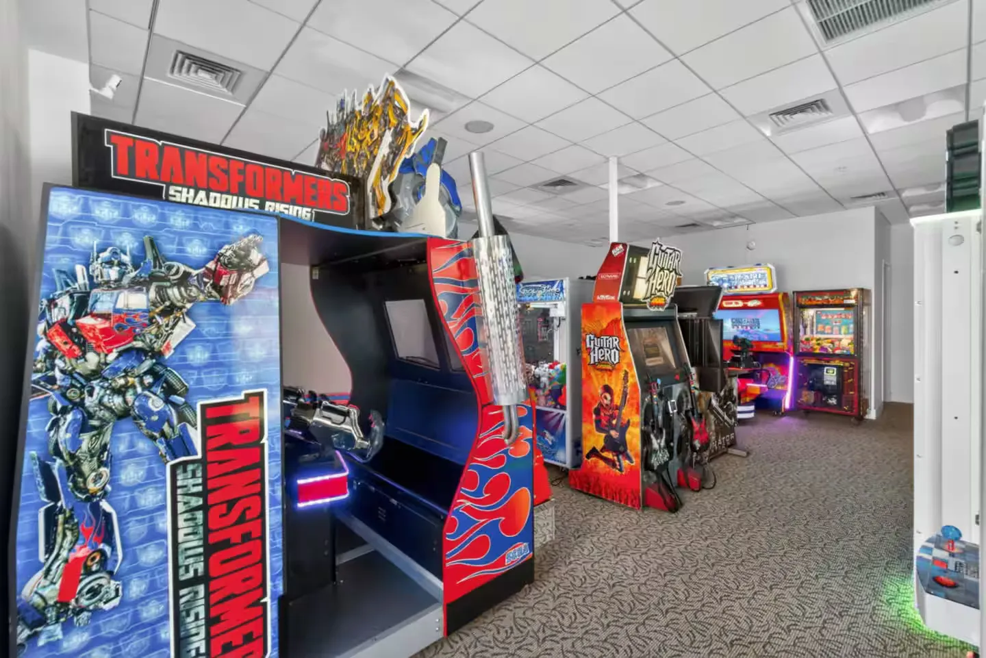 Game room featuring multiple stand-up arcade machines on patterned carpet under a dropped ceiling.