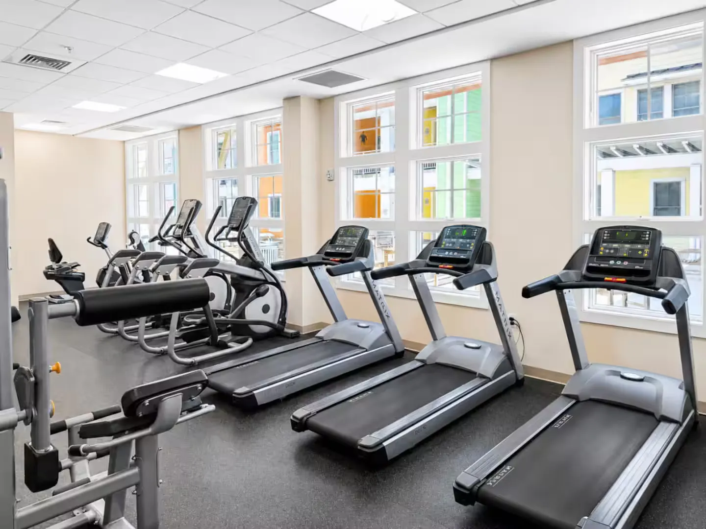 Bright fitness center lined with treadmills, elliptical machines, and exercise equipment beside large windows.