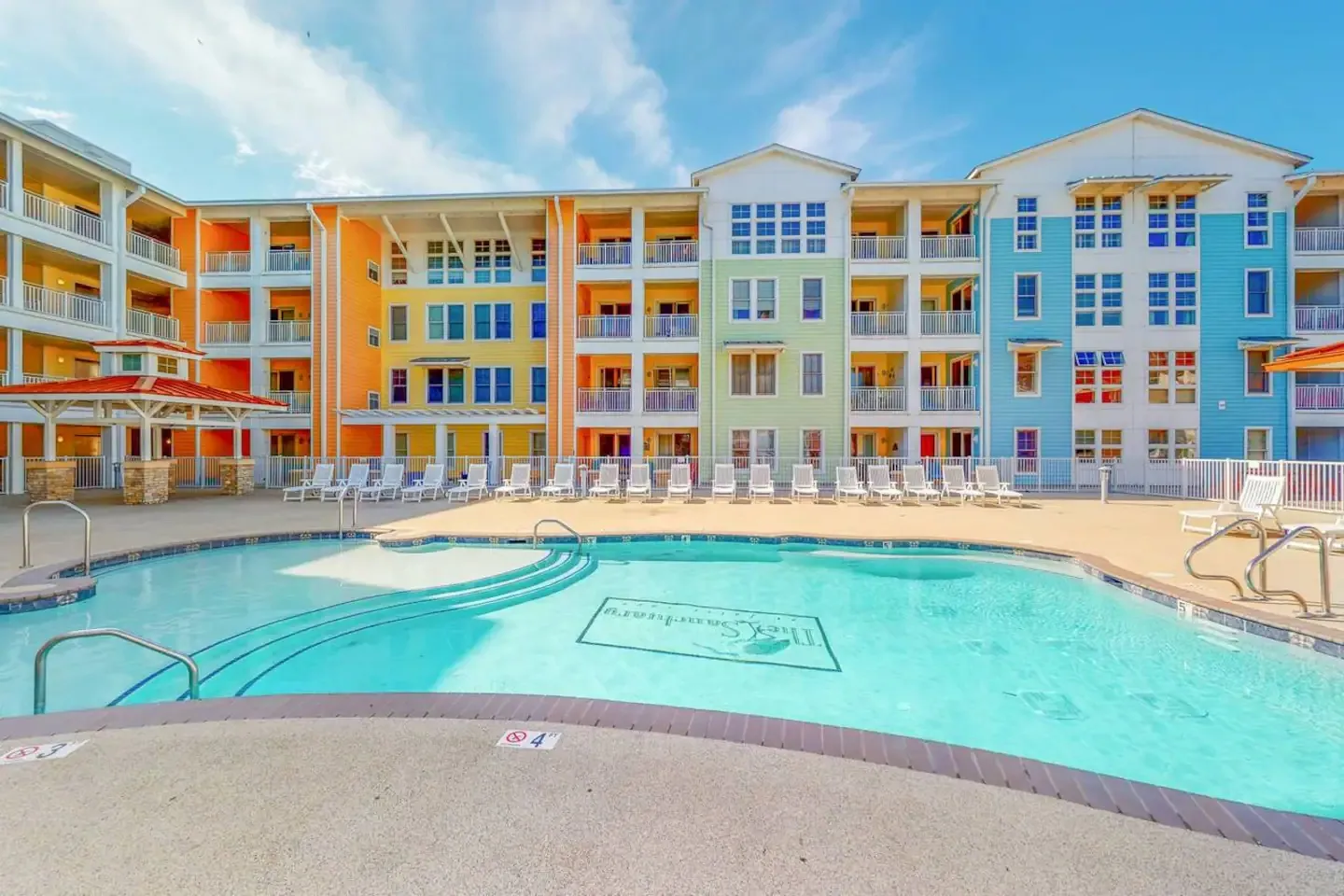 Outdoor swimming pool with lounge chairs in front of a colorful multi-story resort building under a clear blue sky.